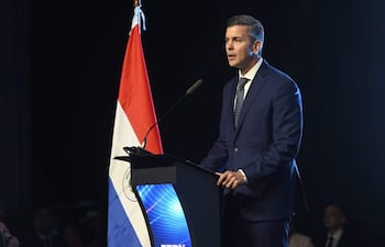 Santiago Peña, con traje oscuro y camisa blanca, habla desde un atril frente a una bandera de Paraguay en un auditorio.
