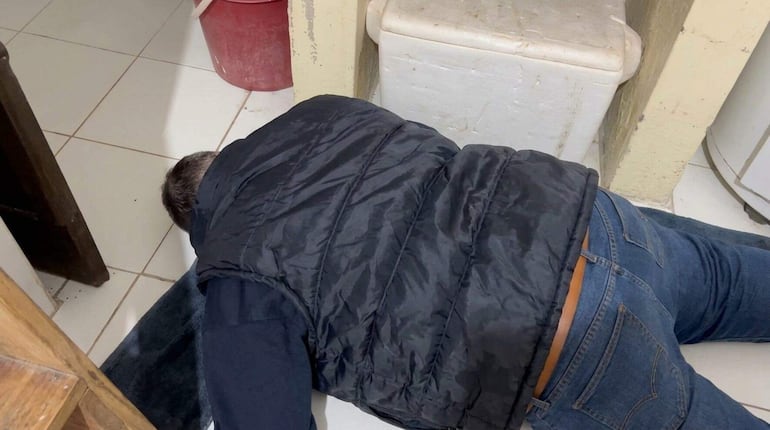 Hombre en chaleco negro y jeans azules, tendido boca abajo en una cocina desordenada.