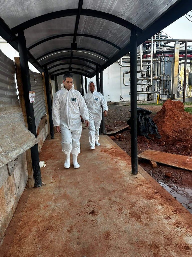 Dos trabajadores con trajes de protección blanca caminan con expresiones serias en un pasillo al aire libre en un entorno industrial.