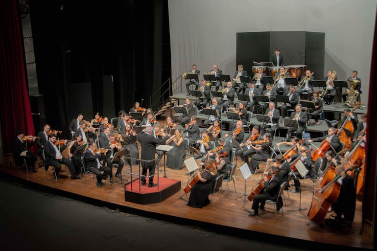 Director de orquesta en traje negro dirige a 40 músicos, todos vestidos formalmente, en ambiente oscuro y con iluminación centrada.