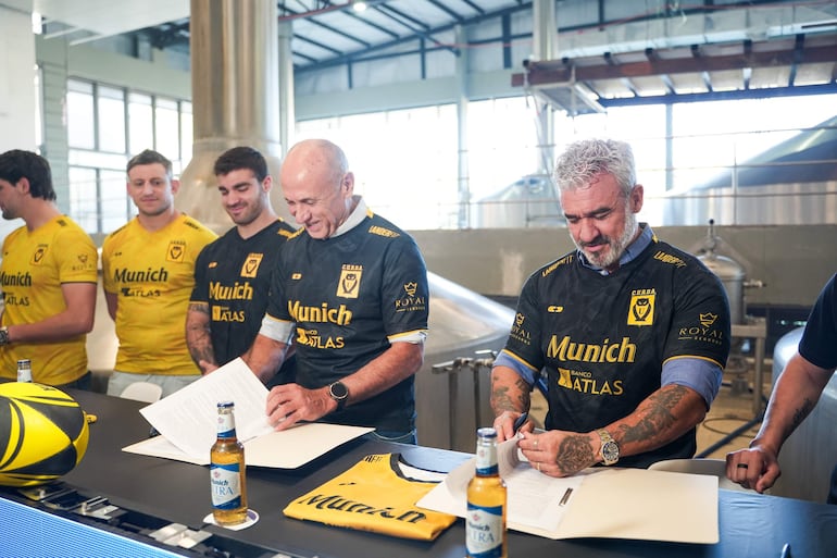 Cinco hombres visten camisetas negras y amarillas de Munich Rugby, organizados tras una mesa con botellas de cerveza y un balón.