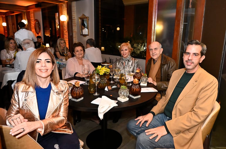 Mujer con chaqueta metálica sonríe, acompañada de otras personas conversando y disfrutando en un ambiente acogedor.
