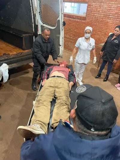 Un hombre de chaqueta oscura atiende a otro en camilla con camiseta roja, rodeados por dos mujeres en un ambiente serio.