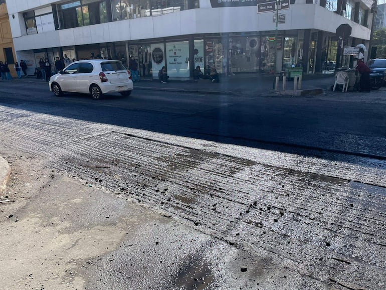 Personas sentadas en la vereda de una calle en reparación, con un auto blanco estacionado y edificios comerciales de fondo.