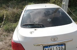 Automóvil Toyota blanco con impactos de bala visible en la luneta, rodeado de pastizales y vegetación.
