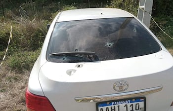 Automóvil Toyota blanco con impactos de bala visible en la luneta, rodeado de pastizales y vegetación.