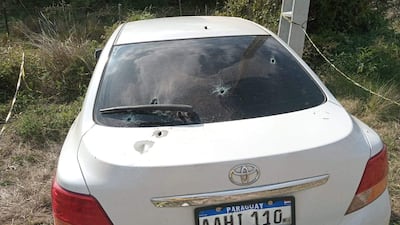 Automóvil Toyota blanco con impactos de bala visible en la luneta, rodeado de pastizales y vegetación.