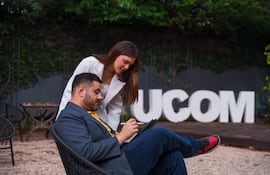Hombre en traje gris y mujer con prenda blanca observan una tablet sentados en sillas bajo un ambiente tranquilo con letrero de UCOM.