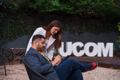 Hombre en traje gris y mujer con prenda blanca observan una tablet sentados en sillas bajo un ambiente tranquilo con letrero de UCOM.