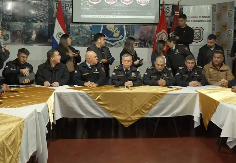 Varios miembros de la Policía Nacional en uniforme oscuro participan en conferencia, escuchando atentamente al orador principal en un salón decorado con banderas.