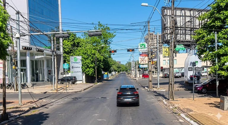 Calle despejada con un automóvil negro en movimiento y otro rojo estacionado, rodeada de árboles y edificios.