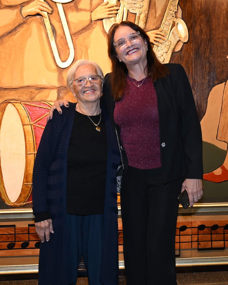 Mujeres sonriendo, una con cabello canoso y blusa negra y otra con cabello rojo y blusa morada, frente a un mural musical.