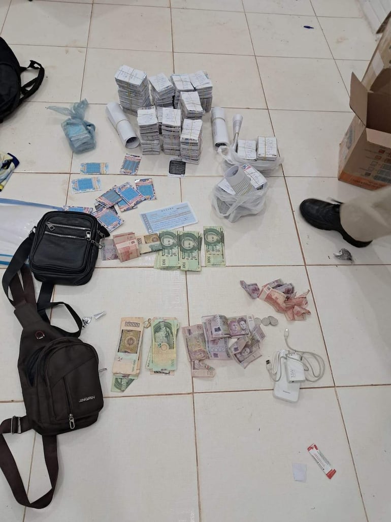 Pilas de billetes y documentos esparcidos en el suelo, junto a una mochila negra y otros objetos.