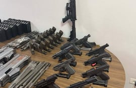 Mesa de madera con diversas armas de fuego, incluyendo pistolas y un rifle de asalto, en un ambiente institucional.