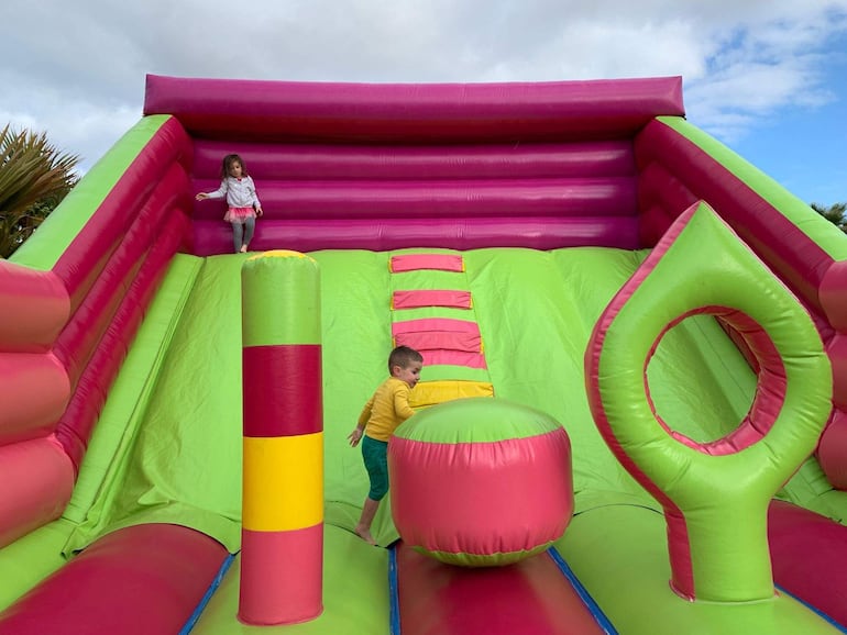 Niña en la parte superior y niño en la parte inferior de un castillo inflable rosa y verde, ambos luciendo felices mientras saltan.