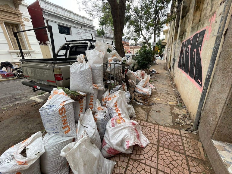 Dos personas frente a un vehículo, manipulando bolsas de basura desordenadas en una acera llena de residuos.