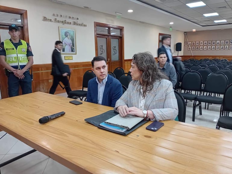 Hernán Rivas en saco azul y camisa blanca, conversa con Alba Zaracho con chaqueta de cuadros, en un auditorio con público vacío.
