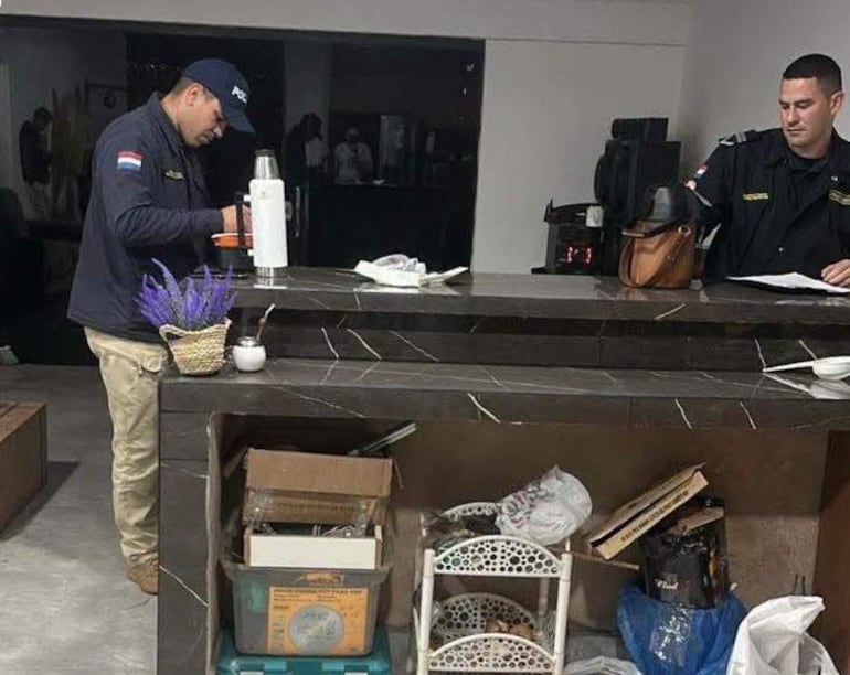Dos hombres en bar, uno tras mostrador con gorro y bandera paraguaya, otro sentado con uniforme observando documentos.