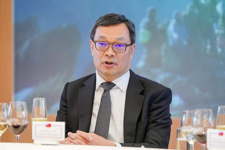 Chen Ming-chi sentado en mesa con traje negro y corbata con puntos, rodeado de copas de vino, en ambiente formal.