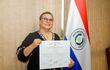 Mujer de cabello corto y gafas, en blusa negra, sonríe mientras sostiene un certificado, frente a una bandera paraguaya.