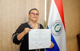 Mujer de cabello corto y gafas, en blusa negra, sonríe mientras sostiene un certificado, frente a una bandera paraguaya.