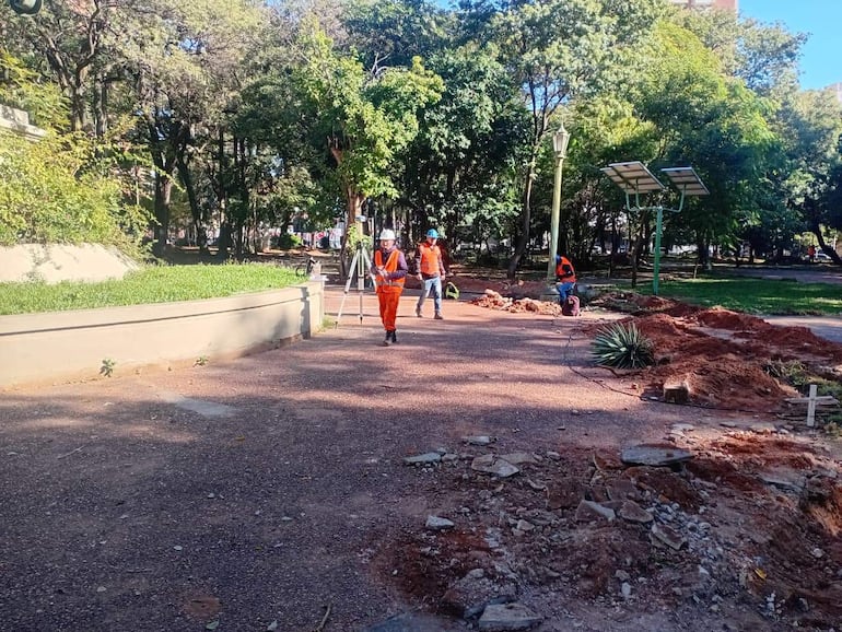 Cuatro personas con chalecos naranjas en un parque en construcción, una mide el terreno con un instrumento mientras dos observan.
