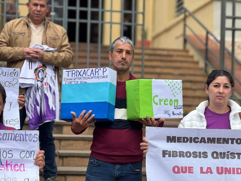 Hombre con cabello corto sostiene una caja de Trixacar, rodeado de mujeres sosteniendo carteles sobre medicamentos necesarios en protesta pacífica.