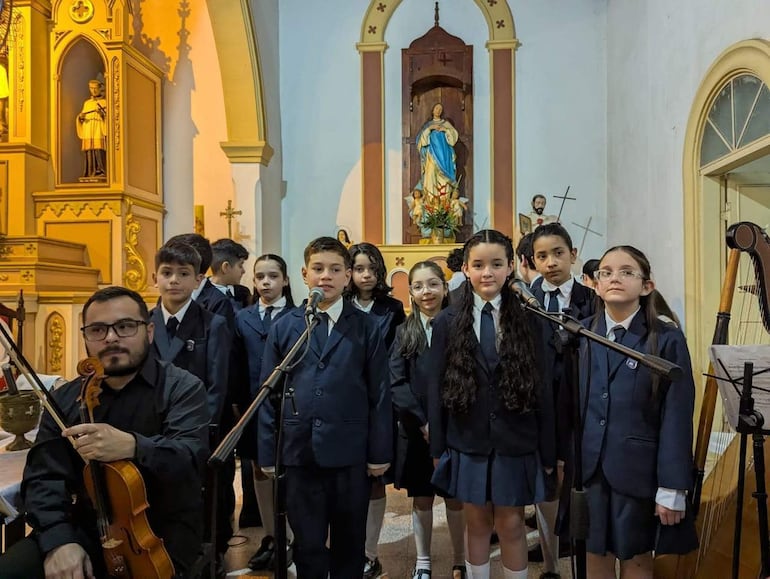 Niños vestidos con uniformes oscuros esperan frente a un micrófono mientras un adulto con violín los acompaña.