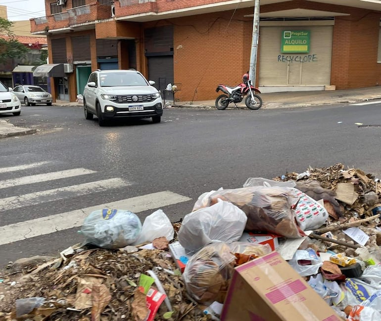 Montículo de basura en la calle con edificios en estado regular, incluyendo vehículos estacionados.