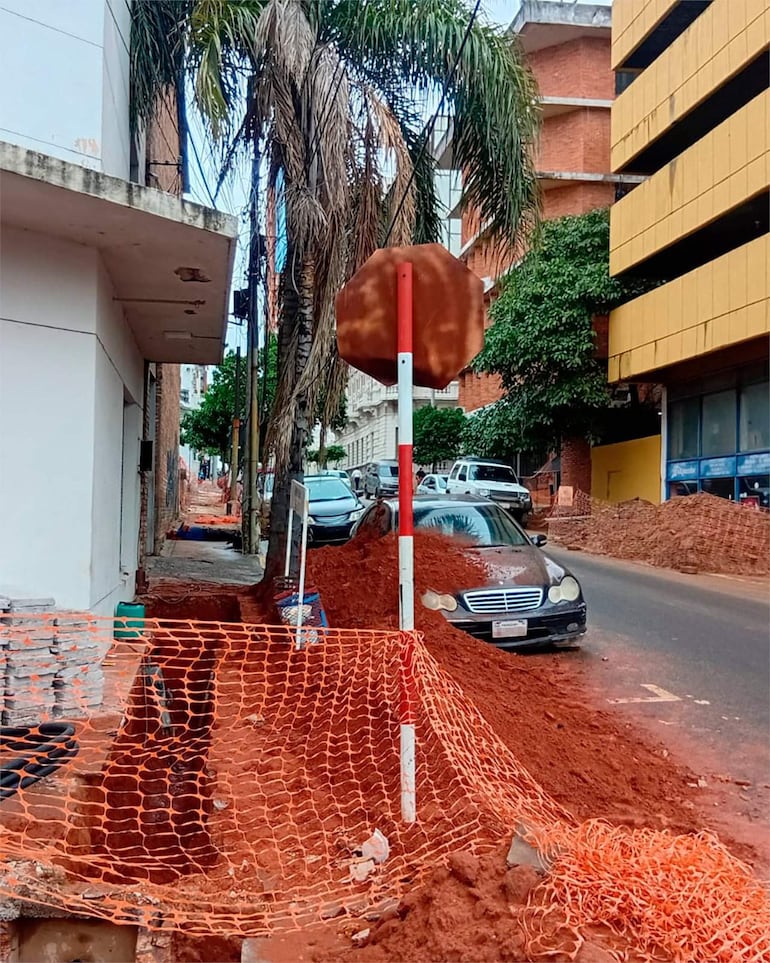 Un auto negro cubierto de tierra roja en una calle urbana, junto a un señalamiento de alto en mal estado y una valla de seguridad.