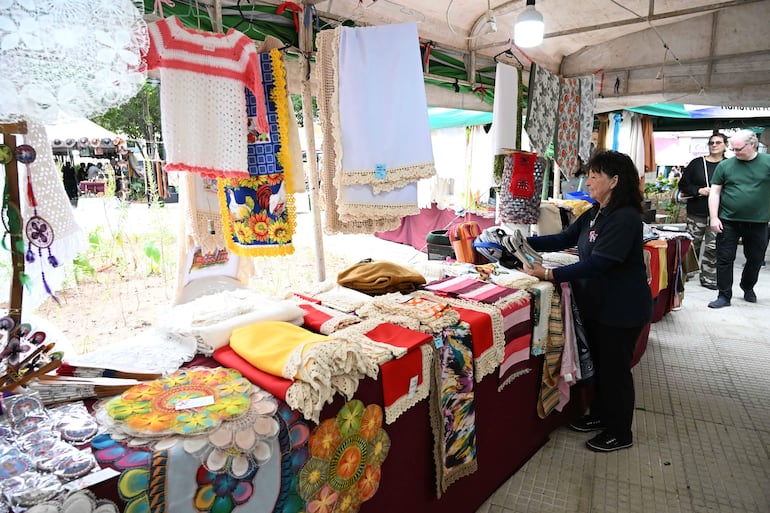 Mujer revisa artesanías sobre mesa, con manteles coloridos; dos hombres observan en un mercado iluminado por luces colgantes.