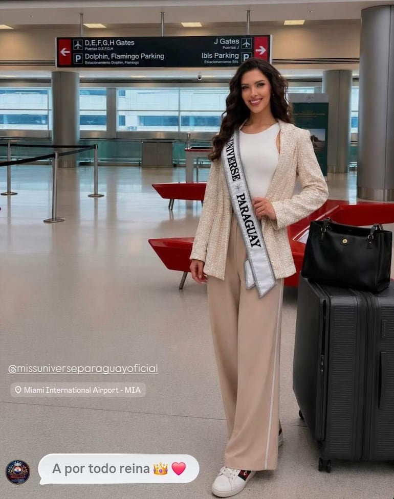 Gretha Matiauda con chaqueta clara y camiseta blanca, sonriente junto a su maleta en el aeropuerto, con rótulos de embarque detrás.