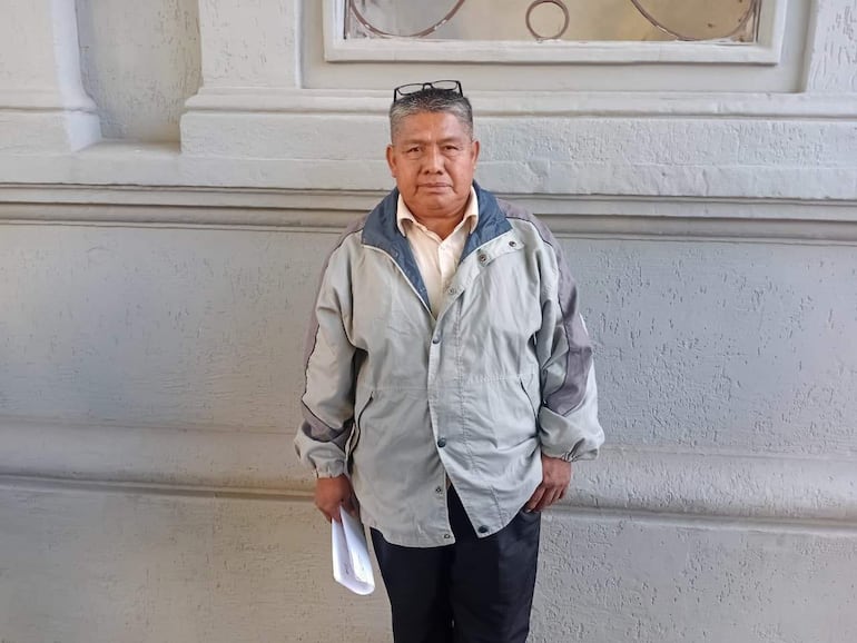 Hombre mayor con chaqueta gris claro y gafas, sosteniendo papeles frente a una pared clara, en ambiente urbano.