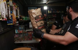 Agente en uniforme y guantes sostiene paquete de 'CARBON' en kisko. Estanterías con productos al fondo.