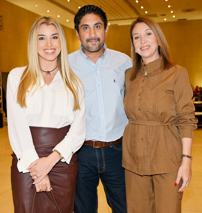 Mujer con blusa blanca y falda marrón, hombre con camisa a rayas, y mujer en blusa marrón, sonríen en un ambiente de celebración.