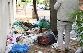 Hombre de espaldas en abrigo claro observa una pila de basura en una calle urbana, con residuos plásticos y papeles mezclados.