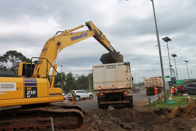 Operario en chaleco naranja supervisa excavadora Komatsu cargando tierra en camión en obra urbana bajo cielo nublado.