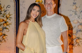 Mujer embarazada con vestido amarillo claro sonríe junto a un hombre en camiseta blanca y pantalones claros, en ambiente festivo.