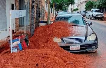Vehículo negro cubierto de tierra roja en la vía pública, con edificios y vegetación de fondo, en un entorno urbano.