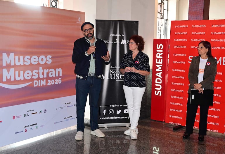 Hombre con boina y gafas en el micrófono, mujer con cabello rizado escucha, otra mujer con carpeta observa en el evento cultural.