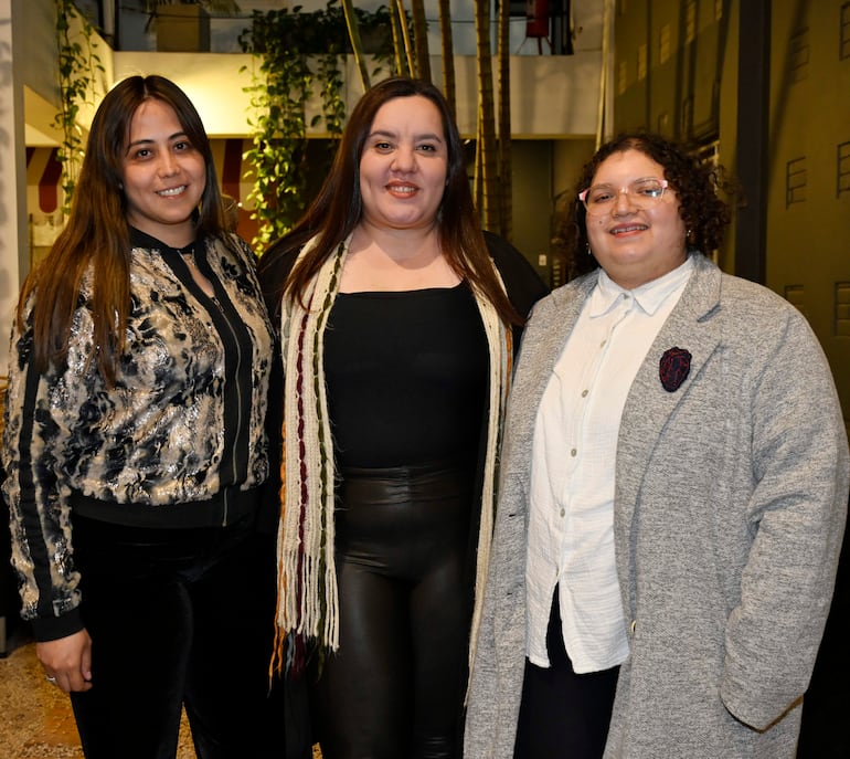Tres mujeres sonrientes: una con chaqueta brillante, otra en top negro y bufanda, y la última con camisa blanca y abrigo gris, en un ambiente decorado.