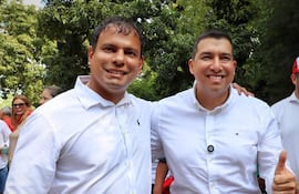 Dos hombres con camisas blancas y sonrisas amplias, uno muestra un pulgar hacia arriba, rodeados de un ambiente natural y personas.