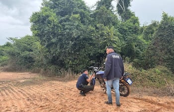 Dos policías, uno agachado cerca de una motocicleta azul, en un entorno boscoso, observando y analizando la escena.