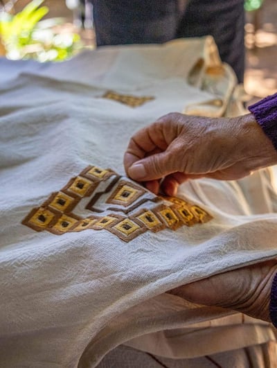 Mano sostiene prenda de tela clara con bordados marrones y dorados, rodeada de vegetación difusa.