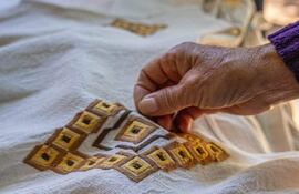 Mano sostiene prenda de tela clara con bordados marrones y dorados, rodeada de vegetación difusa.