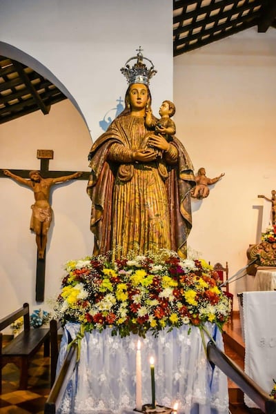 Estatua mariana con niño en brazos adornada con corona, acompañada de un colorido arreglo floral y una cruz en un ambiente de culto.