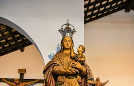 Estatua mariana con niño en brazos adornada con corona, acompañada de un colorido arreglo floral y una cruz en un ambiente de culto.