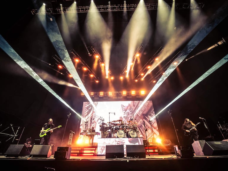 Músicos en escenario iluminado: bajista con bajo verde, tecladista y guitarrista rodeados de luces y efectos láser.