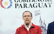 Óscar González, con abrigo rojo y gafas, frente a un micrófono serio, en un ambiente oficial con el emblema del Gobierno del Paraguay detrás.