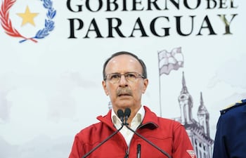 Óscar González, con abrigo rojo y gafas, frente a un micrófono serio, en un ambiente oficial con el emblema del Gobierno del Paraguay detrás.
